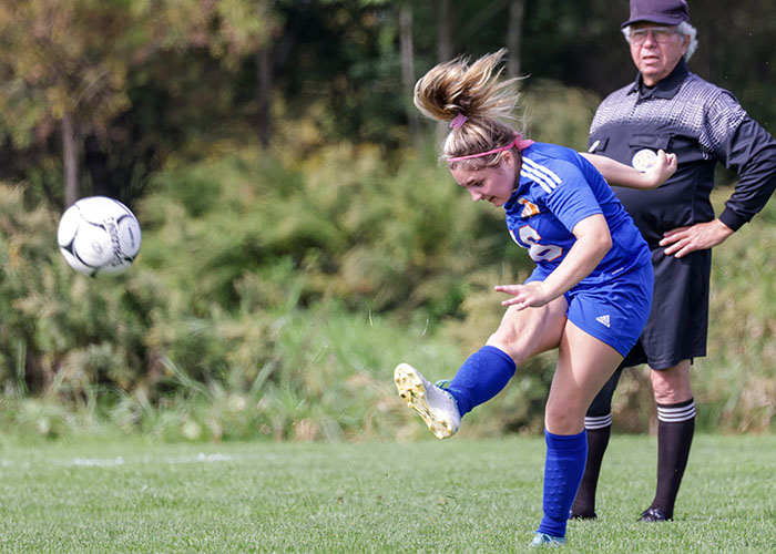 A soccer player kicks a ball as a ref watches