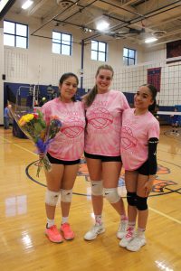 Three volleyball players in pink shirts, one holding flowers, pose