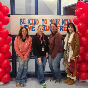 Four people pose in front of a banner flanked by red balloons that reads be kind to your mind live drug free.