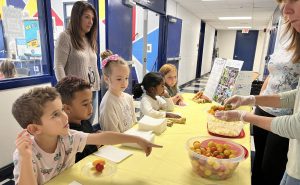 A student points to a bowl of fruit as other students sit at a table with adults behind them.