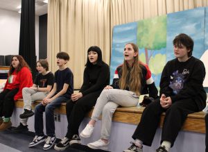 Students sit on the edge of the stage singing during rehearsal for the play