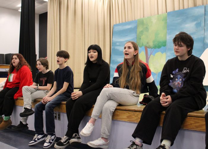 Students sit on the edge of the stage singing during rehearsal for the play